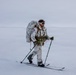 A military personnel in white winter camouflage gear is cross-country skiing across a flat, snow-covered arctic or tundra landscape. The individual is fully equipped with a tactical chest rig, large rucksack, ski poles with green tips, and protective goggles. The overcast, featureless white terrain suggests extreme cold-weather military training or operations.