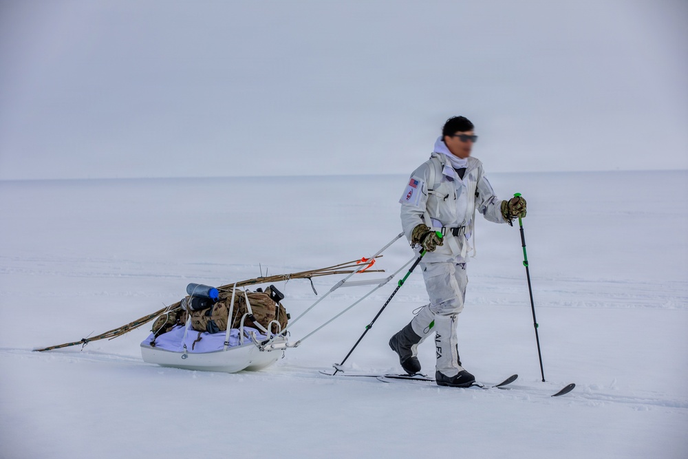 A person in white winter camouflage gear is cross-country skiing across a flat, snow-covered terrain while pulling a loaded pulk (sled) behind them. The individual appears to be on a military or expedition-style arctic traverse, equipped with ski poles, skis branded 'ALFA', and a sled carrying gear including what appears to be a backpack and wooden poles. The environment is a featureless, overcast arctic or polar landscape.