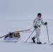 A person in white winter camouflage gear is cross-country skiing across a flat, snow-covered terrain while pulling a loaded pulk (sled) behind them. The individual appears to be on a military or expedition-style arctic traverse, equipped with ski poles, skis branded 'ALFA', and a sled carrying gear including what appears to be a backpack and wooden poles. The environment is a featureless, overcast arctic or polar landscape.