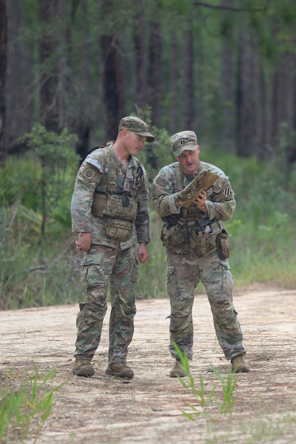 Two U.S. Army soldiers in MultiCam (OCP) camouflage uniforms are standing on a sandy dirt trail in a forested area, reviewing what appears to be a map or document. The soldier on the right is holding and examining the item while appearing to brief or discuss it with the soldier on the left. Both wear tactical vests and patrol caps, and unit patches are visible on their shoulders.