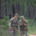 Two U.S. Army soldiers in MultiCam (OCP) camouflage uniforms are standing on a sandy dirt trail in a forested area, reviewing what appears to be a map or document. The soldier on the right is holding and examining the item while appearing to brief or discuss it with the soldier on the left. Both wear tactical vests and patrol caps, and unit patches are visible on their shoulders.