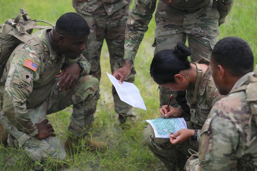 A group of U.S. Army soldiers in Operational Camouflage Pattern (OCP) uniforms are gathered in a grassy field conducting a land navigation exercise. One soldier is actively marking or plotting coordinates on a topographic map while referencing a printed document, with fellow soldiers observing closely. The name tape 'CARBY' is visible on one soldier's uniform, and a U.S. flag patch is visible on their left shoulder.