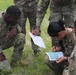 A group of U.S. Army soldiers in Operational Camouflage Pattern (OCP) uniforms are gathered in a grassy field conducting a land navigation exercise. One soldier is actively marking or plotting coordinates on a topographic map while referencing a printed document, with fellow soldiers observing closely. The name tape 'CARBY' is visible on one soldier's uniform, and a U.S. flag patch is visible on their left shoulder.