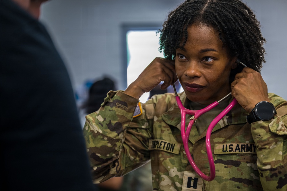 A U.S. Army soldier in OCP (Operational Camouflage Pattern) uniform is placing a pink stethoscope into her ears, preparing to conduct a medical examination. Her name tape reads 'TAPLETON' and her rank insignia indicates she is a Captain (O-3). She appears focused and is facing another individual who is partially visible in the foreground.