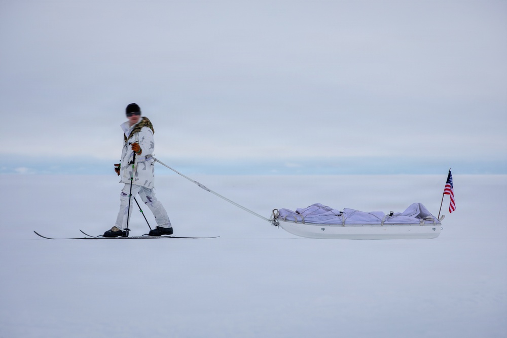 A person dressed in white winter camouflage gear is skiing across a vast, flat snow-covered landscape while pulling a white pulk sled loaded with gear. An American flag is mounted on the rear of the sled. The overcast sky and featureless white terrain suggest an Arctic or Antarctic environment.