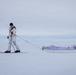 A person dressed in white winter camouflage gear is skiing across a vast, flat snow-covered landscape while pulling a white pulk sled loaded with gear. An American flag is mounted on the rear of the sled. The overcast sky and featureless white terrain suggest an Arctic or Antarctic environment.