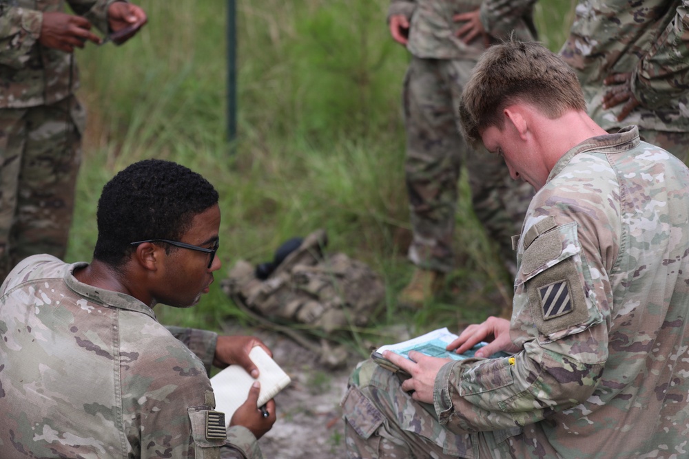 Two U.S. Army soldiers in OCP (Operational Camouflage Pattern) uniforms are seated outdoors, reviewing documents or field manuals together. A shoulder patch on the right soldier's uniform identifies him as belonging to the 3rd Infantry Division. Additional soldiers are visible standing in the background in a grassy, outdoor training environment.