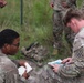 Two U.S. Army soldiers in OCP (Operational Camouflage Pattern) uniforms are seated outdoors, reviewing documents or field manuals together. A shoulder patch on the right soldier's uniform identifies him as belonging to the 3rd Infantry Division. Additional soldiers are visible standing in the background in a grassy, outdoor training environment.