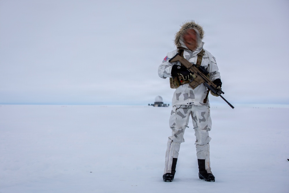 A soldier in white winter camouflage gear stands on a snow-covered terrain, holding an assault rifle. The individual wears a fur-trimmed hood, tactical vest, and black boots, with a US flag patch visible on the left sleeve. A structure consistent with a radar dome or military installation is visible in the distant background.