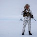 A soldier in white winter camouflage gear stands on a snow-covered terrain, holding an assault rifle. The individual wears a fur-trimmed hood, tactical vest, and black boots, with a US flag patch visible on the left sleeve. A structure consistent with a radar dome or military installation is visible in the distant background.