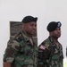 Two military personnel in U.S. Army Battle Dress Uniforms (BDU) woodland camouflage pattern are standing at attention or parade rest near a commemorative plaque on a building wall. Both wear black berets; the soldier on the right has a blue beret flash with a visible insignia patch. The setting appears to be an outdoor military installation ceremony or formal event.