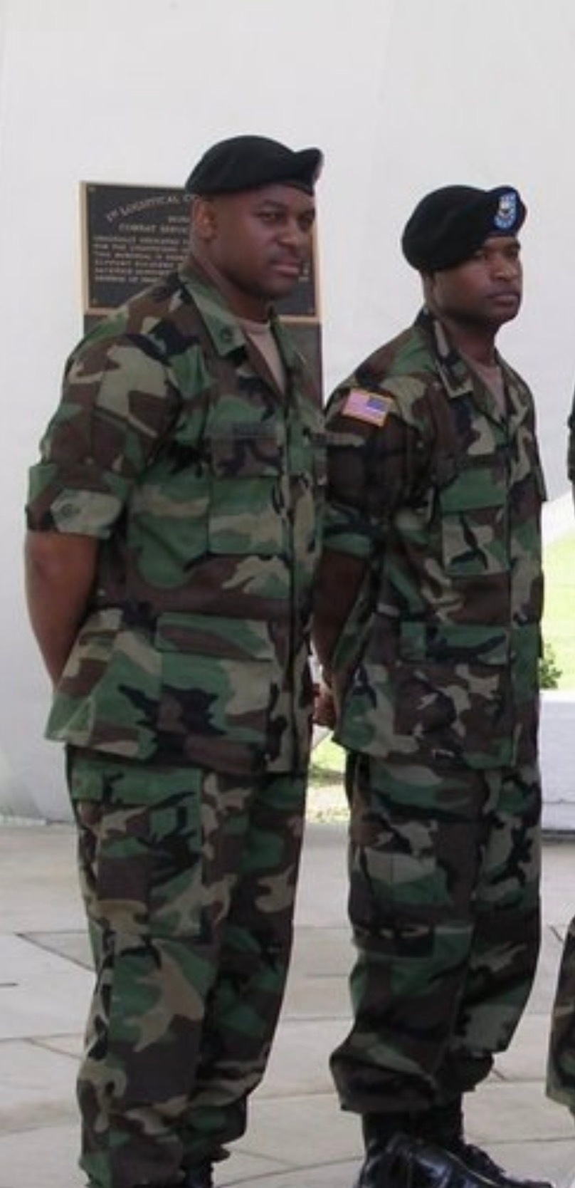 Two military personnel in U.S. Army Battle Dress Uniforms (BDU) woodland camouflage pattern are standing at attention or parade rest near a commemorative plaque on a building wall. Both wear black berets; the soldier on the right has a blue beret flash with a visible insignia patch. The setting appears to be an outdoor military installation ceremony or formal event.