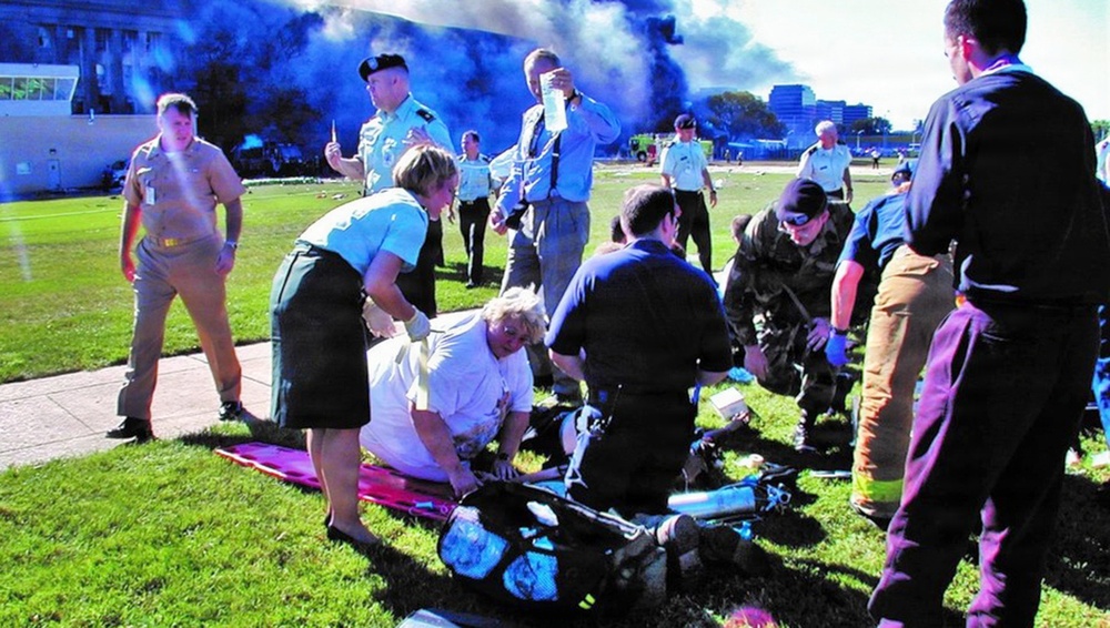 Emergency responders and bystanders are providing medical assistance to injured individuals on a grass lawn. Dark smoke is visible rising in the background near a large building. Multiple personnel in various uniforms are coordinating triage and first aid efforts.