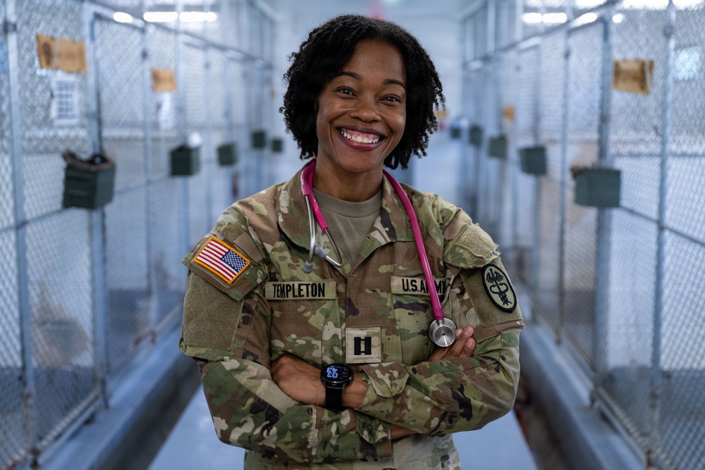 A U.S. Army medical officer named Templeton poses confidently with arms crossed in what appears to be a military kennel or detention facility corridor. She is wearing OCP (Operational Camouflage Pattern) uniform with a pink stethoscope around her neck, indicating a medical role. Her broad smile and relaxed posture suggest a professional portrait or publicity photograph.