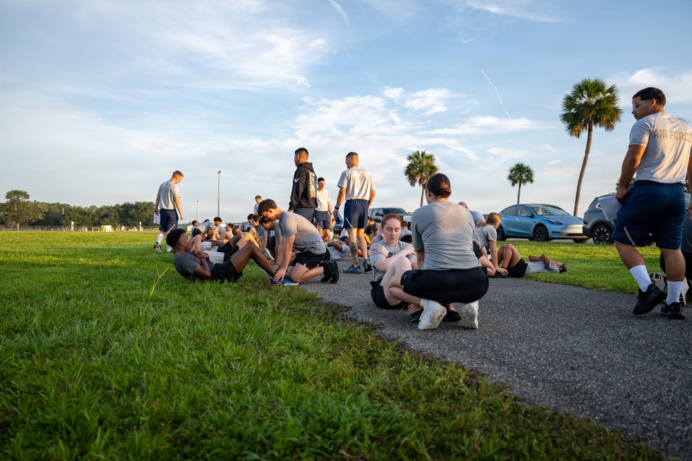 A group of individuals wearing matching gray t-shirts and dark shorts, consistent with Air Force physical training (PT) uniforms, are engaged in outdoor exercise on a paved path and grassy area. The setting appears to be a military base or training facility, identifiable by the 'Air Force' text visible on one shirt. The scene is captured during early morning or late afternoon based on the warm, low-angle sunlight.