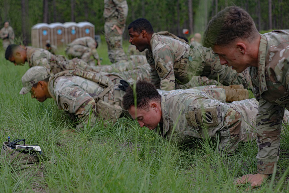 A group of military soldiers in camouflage uniforms are performing push-ups or a low crawl exercise on a grassy field. The soldiers are wearing OCP (Operational Camouflage Pattern) uniforms with unit patches visible on their shoulders, consistent with U.S. Army personnel. The setting appears to be an outdoor training area with portable toilets and wooded terrain visible in the background.