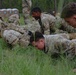 A group of military soldiers in camouflage uniforms are performing push-ups or a low crawl exercise on a grassy field. The soldiers are wearing OCP (Operational Camouflage Pattern) uniforms with unit patches visible on their shoulders, consistent with U.S. Army personnel. The setting appears to be an outdoor training area with portable toilets and wooded terrain visible in the background.