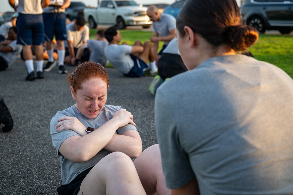 A group of people in gray t-shirts are performing sit-ups on a paved surface, likely during a physical fitness training session. The foreground shows a red-haired woman straining with effort while doing a sit-up, with a partner holding her feet. Multiple participants are visible in the background performing the same exercise near parked vehicles.