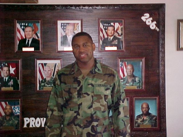 A U.S. Army soldier in woodland camouflage BDU (Battle Dress Uniform) stands in front of a military unit command display board. The board features framed photographs of what appear to be senior military and civilian officials, with the number '266' visible in the upper right corner and partial text 'PROV' visible at the bottom left. The display is mounted on a dark wood panel and likely represents a chain of command or commanders' history board.