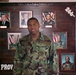 A U.S. Army soldier in woodland camouflage BDU (Battle Dress Uniform) stands in front of a military unit command display board. The board features framed photographs of what appear to be senior military and civilian officials, with the number '266' visible in the upper right corner and partial text 'PROV' visible at the bottom left. The display is mounted on a dark wood panel and likely represents a chain of command or commanders' history board.