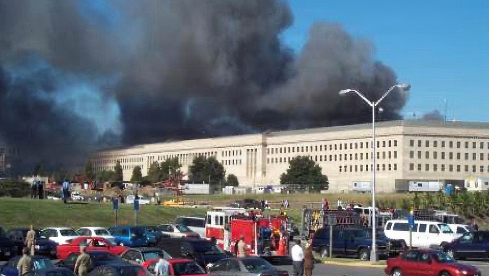 The image shows the Pentagon building in Arlington, Virginia with massive black smoke rising from its structure, consistent with the September 11, 2001 terrorist attack. Emergency response vehicles, including fire trucks, are visible in the foreground parking area along with numerous personnel. The scene depicts an active emergency response to a large-scale structural fire or explosion.