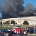 The image shows the Pentagon building in Arlington, Virginia with massive black smoke rising from its structure, consistent with the September 11, 2001 terrorist attack. Emergency response vehicles, including fire trucks, are visible in the foreground parking area along with numerous personnel. The scene depicts an active emergency response to a large-scale structural fire or explosion.