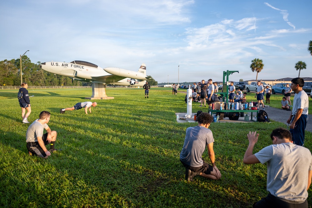 A group of individuals in athletic/military PT gear are conducting physical training exercises on a grass field at what appears to be a U.S. Air Force base. A static display aircraft marked 'U.S. AIR FORCE' with tail number 5325 is mounted on a pedestal in the background. The scene appears to be an early morning fitness session with various participants at different stages of exercise.
