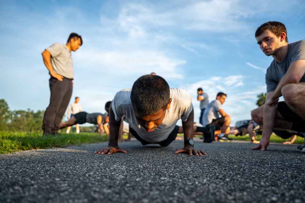 A group of individuals are performing physical fitness training outdoors on a paved surface. The foreground shows one person doing push-ups at ground level, while others are visible in the background doing similar exercises. One person appears to be observing or supervising the activity.