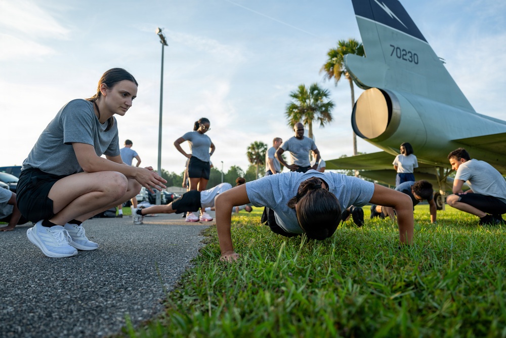 A group of individuals in matching gray t-shirts and dark shorts are performing physical fitness training outdoors near a static display aircraft. One person is doing push-ups on grass while another crouches nearby observing and providing guidance. The setting appears to be a military or ROTC training environment, with a jet aircraft tail section bearing the number '70230' visible in the background.