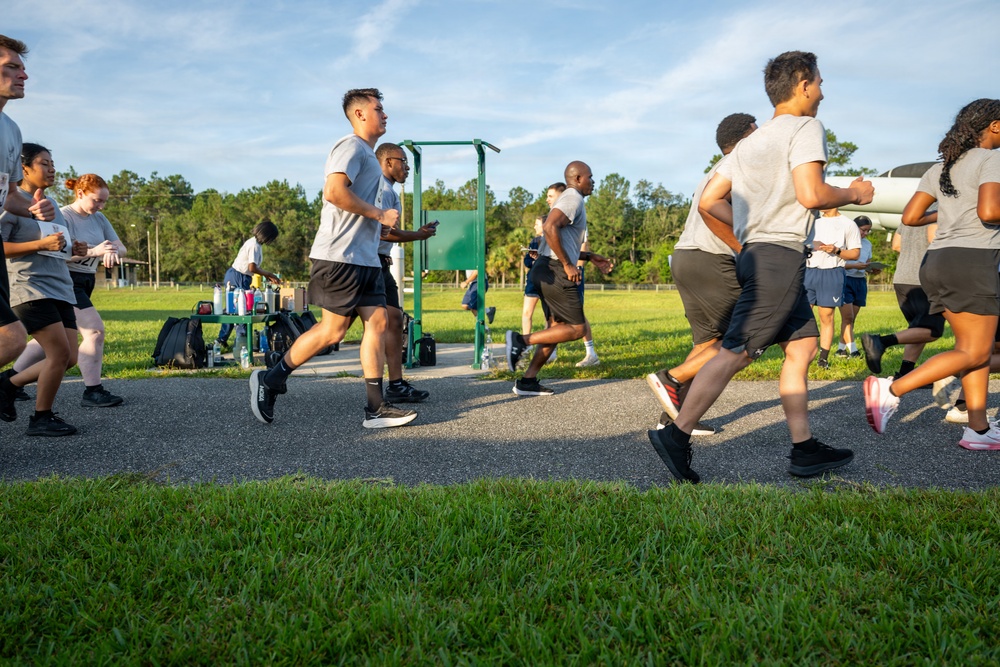 A large group of people in matching gray t-shirts and black shorts are running together on a paved path surrounded by green grass. The setting appears to be an outdoor fitness or training area, likely a military or institutional physical training session. The image is taken from a low angle during what appears to be early morning or late afternoon based on the warm, directional sunlight.