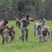 A group of military personnel in camouflage uniforms are conducting physical training or a field exercise in an open grassy area. The soldiers appear to be performing ground-based drills, with some crouching, crawling, or rising from the ground. The setting appears to be a military training ground bordered by a tree line.