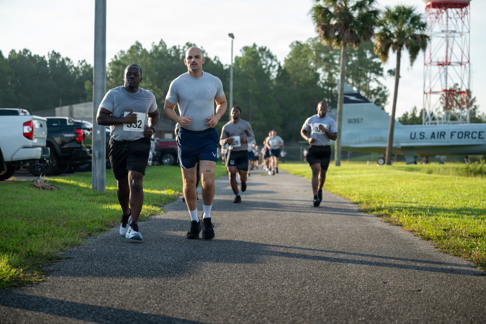 A group of U.S. Air Force personnel are participating in an organized run or fitness event on a paved path on what appears to be a military base. Runners are wearing race bibs with numbers visible, including bib numbers 332 and 32. In the background, a static display aircraft marked 'U.S. AIR FORCE' and a red-and-white communications or radar tower are visible, confirming the military installation setting.
