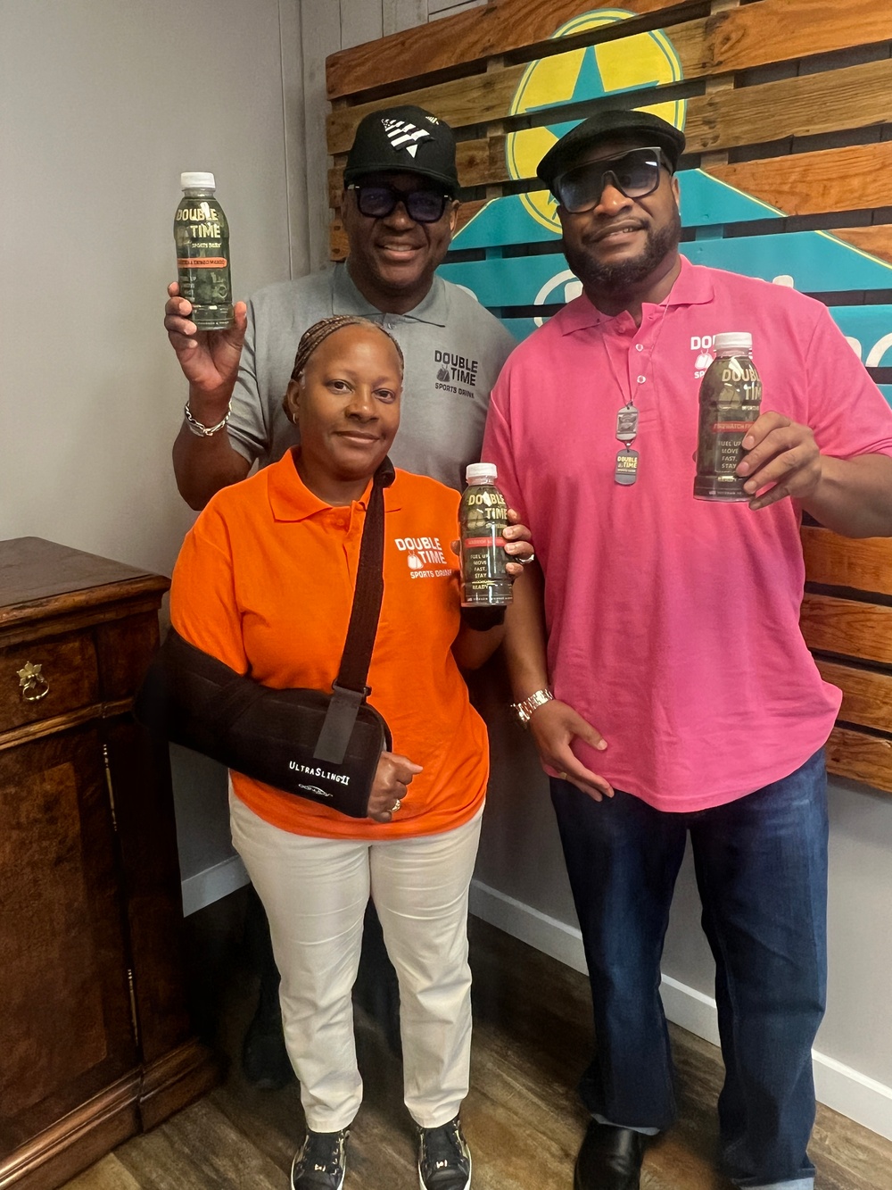 Three individuals are posing together while holding bottles of 'Double Time Sports Drink.' They are wearing branded polo shirts and caps promoting the same product, suggesting they are associated with the brand. The setting appears to be an indoor promotional or retail space with a decorative wooden pallet wall backdrop.