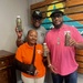 Three individuals are posing together while holding bottles of 'Double Time Sports Drink.' They are wearing branded polo shirts and caps promoting the same product, suggesting they are associated with the brand. The setting appears to be an indoor promotional or retail space with a decorative wooden pallet wall backdrop.