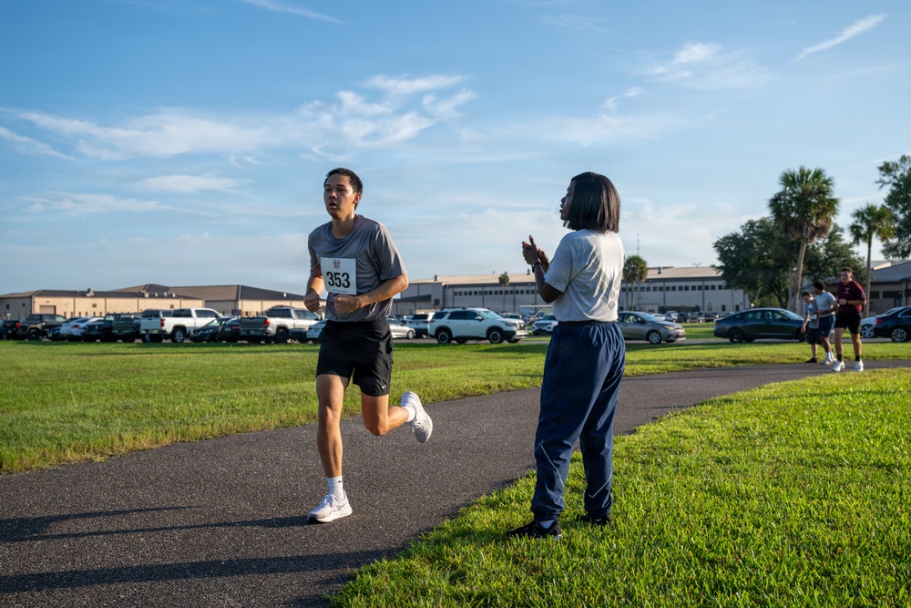 A runner wearing bib number 353 is competing in an outdoor race on a paved path, likely at a military base given the institutional buildings and setting. A person in navy blue athletic pants and a white shirt stands nearby, appearing to clap or cheer for the runner. The scene takes place during morning or late afternoon hours based on the warm, low-angle sunlight.
