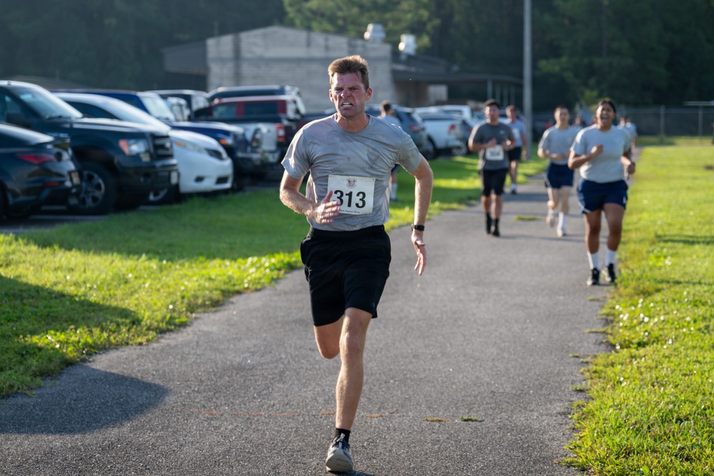 A competitive running race is taking place on a paved path, with bib number 313 leading the pack with an intense, strained expression. Several other runners follow behind on the same path, with a parking lot visible to the left and green grass and trees in the background. The setting appears to be a organized 5K or similar road race at what looks like a military or institutional facility.