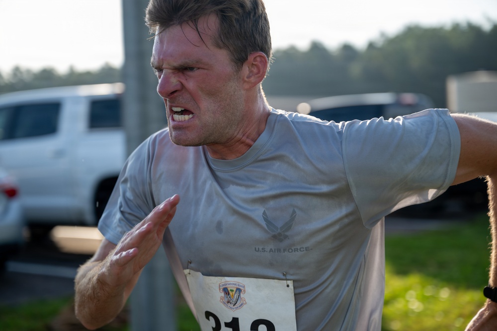 A man wearing a U.S. Air Force t-shirt is competing in a road race, displaying intense physical exertion with a grimacing facial expression. He is wearing race bib number 312 and appears to be pushing hard, likely near the finish line. The background shows parked vehicles and trees in soft focus.