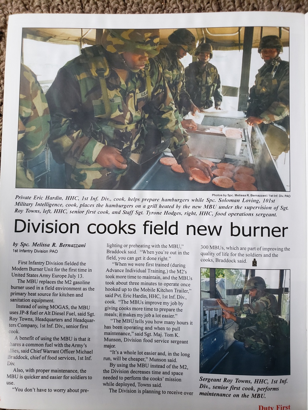 A military newspaper article titled 'Division cooks field new burner' written by Spc. Melissa R. Bernazzani of the 1st Infantry Division PAO. The article covers the First Infantry Division fielding the Modern Burner Unit (MBU) for the first time in United States Army Europe on July 13. Two photographs accompany the article showing soldiers in camouflage uniforms preparing hamburgers on a grill and performing maintenance on the MBU.