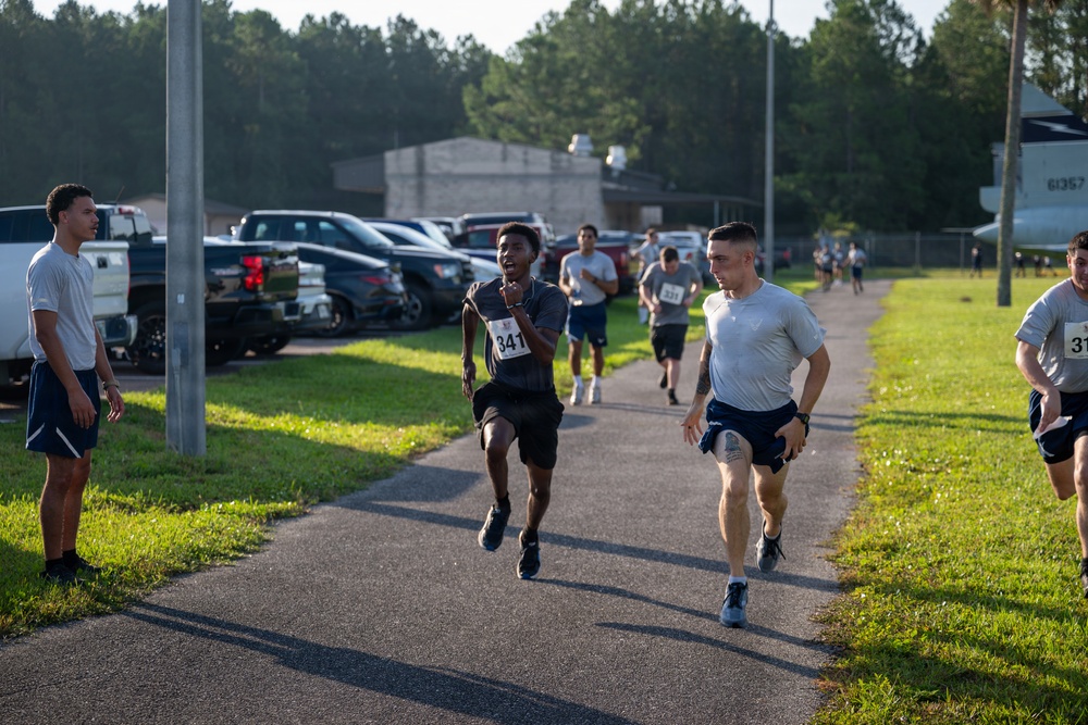 A group of runners participate in an organized race or fitness run event, likely on a military base given the aircraft visible in the background and the institutional setting. The foreground features two lead runners wearing race bibs (numbers 341 and an unidentified number), with numerous other participants visible behind them on a paved path. The event appears to take place in the early morning based on the low-angle sunlight and long shadows.