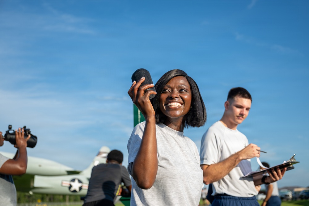 A smiling woman in a light gray t-shirt holds a small cylindrical object (likely a portable speaker or microphone) near her face while looking upward. She is outdoors at what appears to be an airfield or aviation event, with a vintage military aircraft bearing a U.S. star insignia visible in the background. Several other individuals are present, including a man writing on a clipboard and another person using a camera.