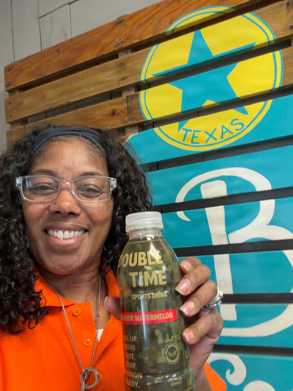 A smiling woman wearing an orange polo shirt and clear-framed glasses holds up a bottle of 'Double Time Sports Drink' in the Watermelon flavor. She is posing in front of a teal banner featuring a yellow star logo with the word 'TEXAS,' mounted on a wooden pallet wall. The image appears to be a promotional or endorsement photo.