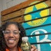 A smiling woman wearing an orange polo shirt and clear-framed glasses holds up a bottle of 'Double Time Sports Drink' in the Watermelon flavor. She is posing in front of a teal banner featuring a yellow star logo with the word 'TEXAS,' mounted on a wooden pallet wall. The image appears to be a promotional or endorsement photo.