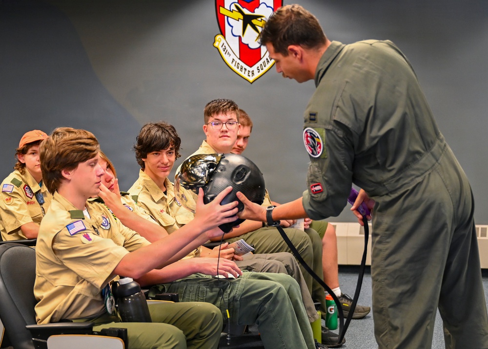 A military pilot in an olive green flight suit is presenting equipment to a group of young scouts seated in a briefing room. The pilot is showing what appears to be a pilot's helmet to the attentive youth group. The setting is an official military squadron briefing room, identifiable by the unit insignia on the wall reading '131st Fighter Squadron.'