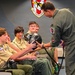 A military pilot in an olive green flight suit is presenting equipment to a group of young scouts seated in a briefing room. The pilot is showing what appears to be a pilot's helmet to the attentive youth group. The setting is an official military squadron briefing room, identifiable by the unit insignia on the wall reading '131st Fighter Squadron.'