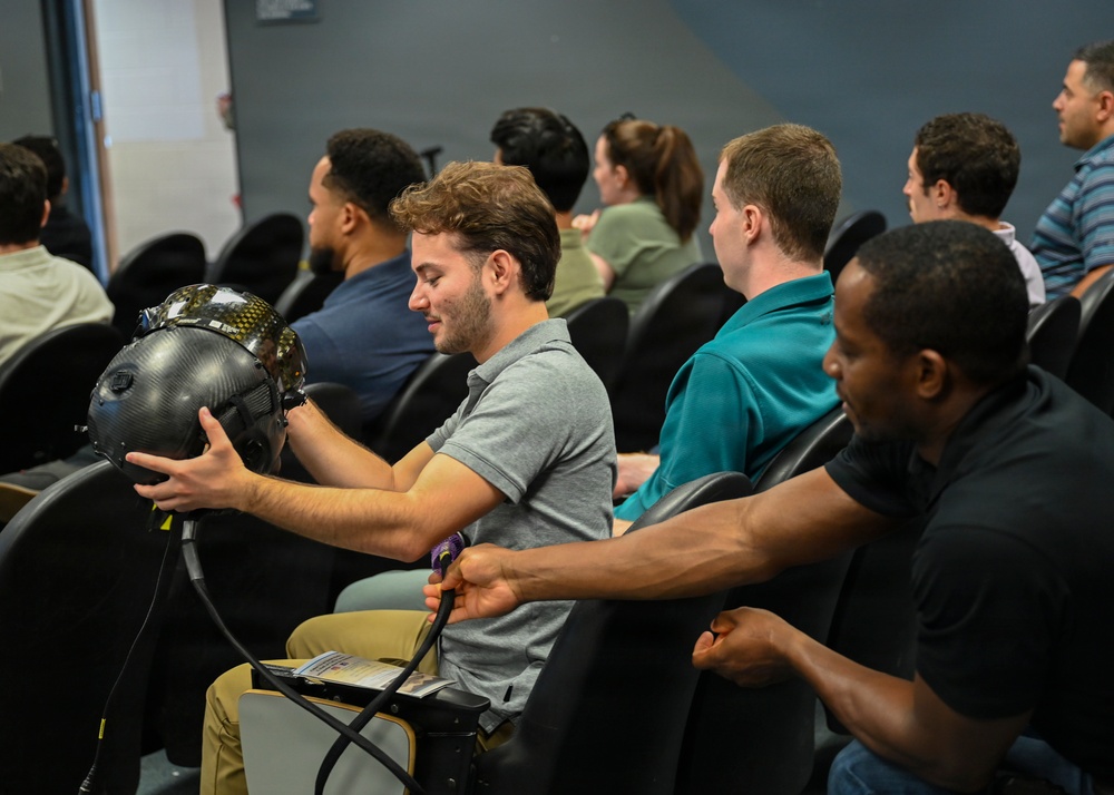 A group of people are seated in a briefing or classroom-style setting with black leather chairs. A young man in the foreground is examining a dark carbon-fiber helmet, which is being passed or handed between attendees. The setting appears to be a professional or training environment, possibly military or aviation-related given the specialized helmet.