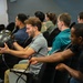A group of people are seated in a briefing or classroom-style setting with black leather chairs. A young man in the foreground is examining a dark carbon-fiber helmet, which is being passed or handed between attendees. The setting appears to be a professional or training environment, possibly military or aviation-related given the specialized helmet.