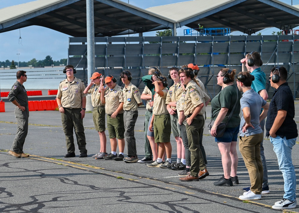 A group of Boy Scouts in tan uniforms and adults are gathered on an airfield tarmac, wearing hearing protection ear muffs. A person in a flight suit on the left appears to be briefing or instructing the group. The setting appears to be a military or civilian airshow or airfield tour event.