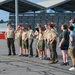A group of Boy Scouts in tan uniforms and adults are gathered on an airfield tarmac, wearing hearing protection ear muffs. A person in a flight suit on the left appears to be briefing or instructing the group. The setting appears to be a military or civilian airshow or airfield tour event.