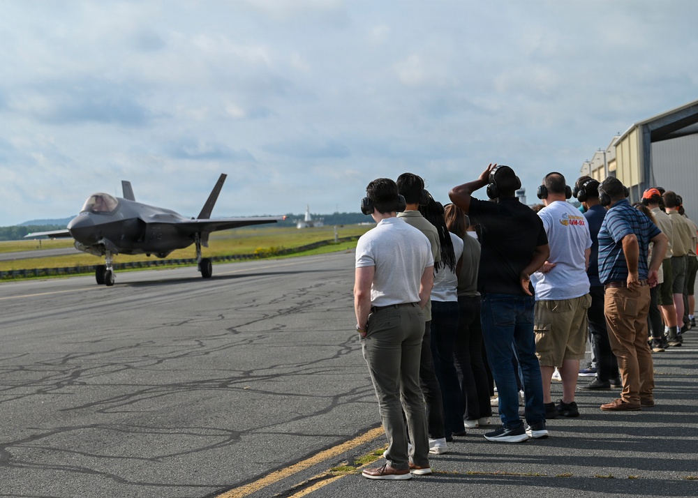 A group of civilians and personnel wearing hearing protection stand along the edge of a military airfield tarmac, observing an F-35 Lightning II stealth fighter jet taxiing on the runway. The setting appears to be an organized visit or demonstration event at a military air base, with hangars visible in the background. The observers are lined up in a row, several shielding their eyes or adjusting their ear protection.