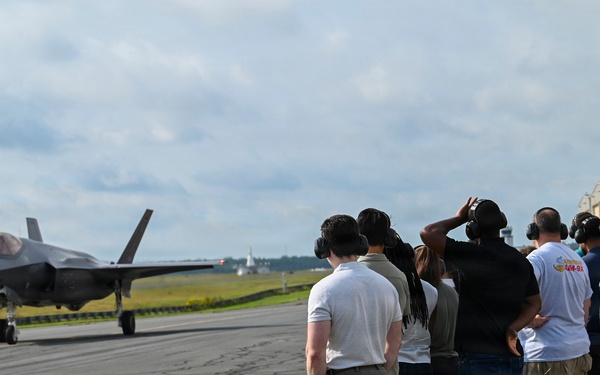 A group of civilians and personnel wearing hearing protection stand along the edge of a military airfield tarmac, observing an F-35 Lightning II stealth fighter jet taxiing on the runway. The setting appears to be an organized visit or demonstration event at a military air base, with hangars visible in the background. The observers are lined up in a row, several shielding their eyes or adjusting their ear protection.